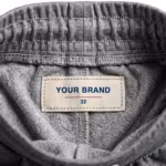 Close-up of the inside back waistband of the men's grey marl interlock knit track pants showing the cream woven YOUR BRAND label with navy lettering, a red rule line and the size 32 mark, stitched into the waistband