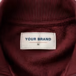 Close-up of the inside back neck of the burgundy tricot tracksuit jacket showing the cream woven YOUR BRAND label with navy lettering, a red rule line and the size M mark, stitched into the inside back neck below the ribbed stand collar